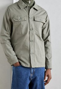 Homme portant une veste kaki boutonnée par-dessus un haut vert zippé avec un jean bleu, debout, une main dans la poche devant un fond uni.