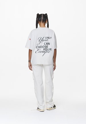 Osoba z warkoczami, ubrana w biały, oversize t-shirt z napisem „Only You Can Choose Your Escape” oraz białe spodnie cargo, stojąca tyłem.