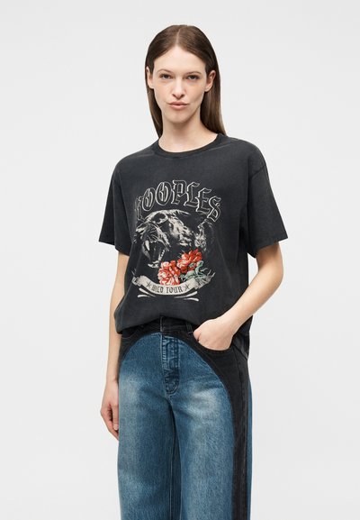 Γυναίκα με μαύρο oversized t-shirt που έχει μια ουρλιάζουσα πάνθηρα, κόκκινα λουλούδια και το κείμενο "The Kooples Wild Tour", συνδυασμένο με δίχρωμα μπλε τζιν.