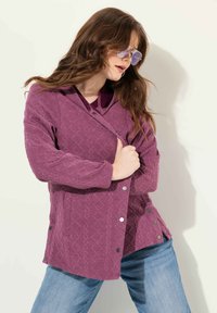 Lila texturerad jacka med ett diamantmönster, har huva, tryckknappar och sidofickor, kombinerad med ljusblå jeans.