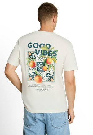 Mężczyzna ubrany w białą koszulkę z napisem "GOOD VIBES", ilustracjami pomarańczy i liścia oraz jasnoniebieskie dżinsy, stojący tyłem do kamery.