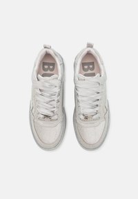 Stříbrné a bílé tenisky s texturovaným síťovým svrškem, hladkými tkaničkami a logem. Mají polstrovanou podrážku a lesklý povrch.