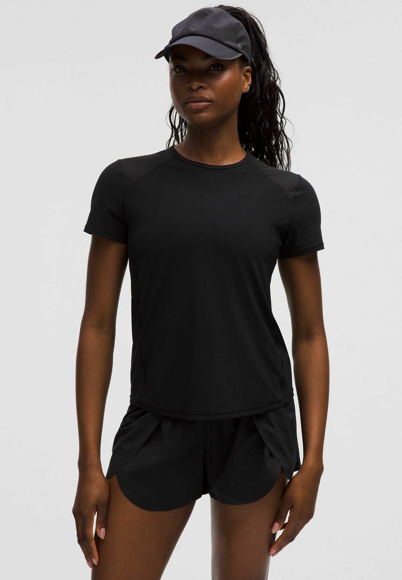 Atletische vrouw in een zwart T-shirt met korte mouwen, zwarte hardloopshorts en een zwarte zonneklep, staande tegen een effen lichte achtergrond.