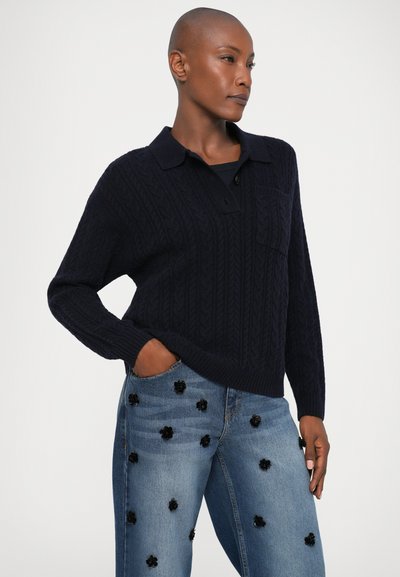 Marineblå kabelstrikket genser med krage og knappestolpe, kombinert med lyseblå denimjeans med svarte blomsterapplikasjoner.