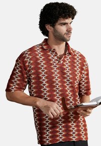 Polo con un patrón en zigzag rojo y blanco, con mangas cortas y un cuello. La tela parece texturizada y tiene un corte holgado.