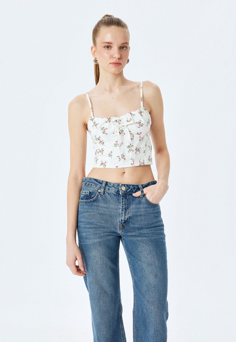 Uttagen vit blommig linne med tunna band, med samlad byst och små rosa och gröna blommönster, parat med högmidjade blå jeans.