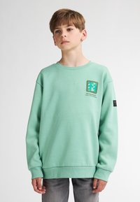Sweatshirt vert menthe avec des poignets et un ourlet côtelés, arborant un logo graphique en carré sur la poitrine et une étiquette noire sur la manche.