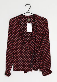 Black blouse with red polka dots, featuring a ruffled neckline, long sleeves, and a tie closure at the waist. Smooth fabric, casual design.
