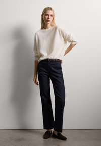 Maglione a maglia color crema, pantaloni in denim blu scuro, cintura in pelle marrone e scarpe slip-on marrone scuro. Vestibilità semplice e rilassata.