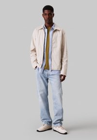 Giacca beige chiaro sopra una camicia verde e una camicia a righe con bottoni, abbinata a jeans azzurri e sneakers bianche, in piedi davanti a uno sfondo grigio.