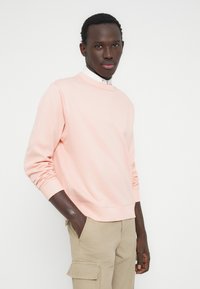 Hombre con sudadera de color rosa claro, camisa blanca debajo y pantalones cargo beige, de pie con una mano en el bolsillo contra un fondo blanco.