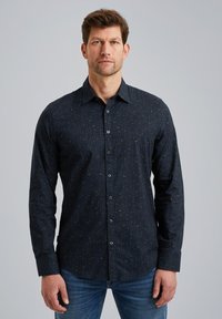 Chemise à manches longues bleu foncé avec un motif subtil, col à boutons, et boutons à l'avant. Tissu doux avec une texture lisse. Jean bleu porté.
