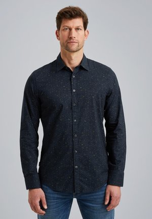 Mørkeblå langærmet skjorte med et diskret mønster, knap-ned krave og knapper foran. Blød stof med en glat tekstur. Blå jeans er på.