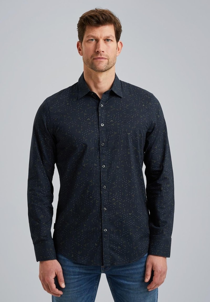 Chemise à manches longues bleu foncé avec un motif subtil, col à boutons, et boutons à l'avant. Tissu doux avec une texture lisse. Jean bleu porté.