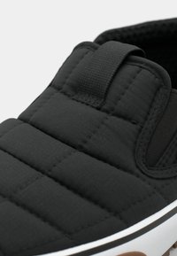 Svart quiltet slip-on sko med teksturert overdel, polstret tekstil og løkke-stropp. Hvit mellomlcollection og brun gummisåle er synlig.