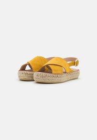 Yellow suede sandals with cutout design, adjustable ankle strap, and woven jute soles. Features smooth leather inner lining.