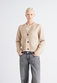 Μπεζ πλεκτό πουλόβερ με V-λαιμό, ραμμένα μανίκια, δύο μπροστινές τσέπες και σκούρα κουμπιά, συνδυασμένο με ανοιχτό γκρί τζιν.