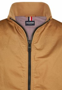 Beige Jacke mit hohem Kragen, ausgestattet mit einem schwarzen Reißverschluss und einem gefütterten Innenraum mit rot-weißen Streifen. Markenlabel: Hechter Paris.
