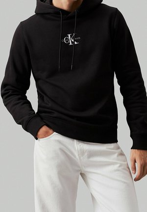 Mann trägt schwarzen Calvin Klein Hoodie und weiße Hose, mit einer Hand in der Tasche vor einfarbig grauem Hintergrund.