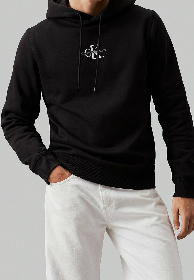 Homme portant un sweat à capuche noir Calvin Klein et un pantalon blanc, avec une main dans la poche, sur fond gris uni.