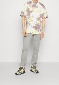 Tie-dye T-shirt i gult och brunt, lös passform; ljusgrå cargobyxor; gröna och svarta sportskor med gula detaljer och bruna snören.