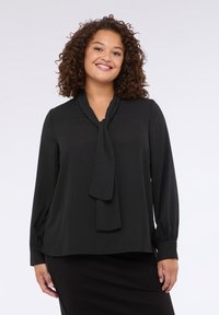 Blusa nera con collo a cravatta, maniche lunghe e un delicato motivo a pois. Tessuto morbido con una vestibilità loose, abbinata a una gonna nera semplice.