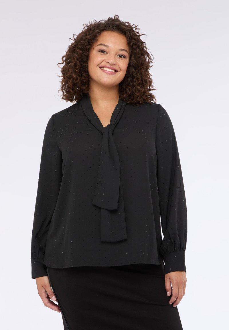 Blusa nera con collo a cravatta, maniche lunghe e un delicato motivo a pois. Tessuto morbido con una vestibilità loose, abbinata a una gonna nera semplice.
