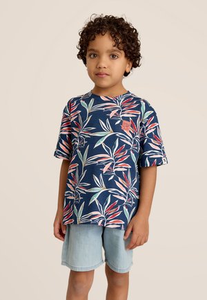 Chemise bleue à manches courtes avec un motif de feuilles tropicales dans des nuances de vert et de rose ; portée avec un short en jean clair. Détail de l'étiquette sur la chemise.