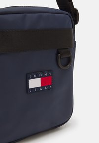 Bolso de nylon azul marino tipo bandolera con una correa ajustable negra, que presenta un parche con logo con acentos en rojo, blanco y azul. Superficie texturizada.