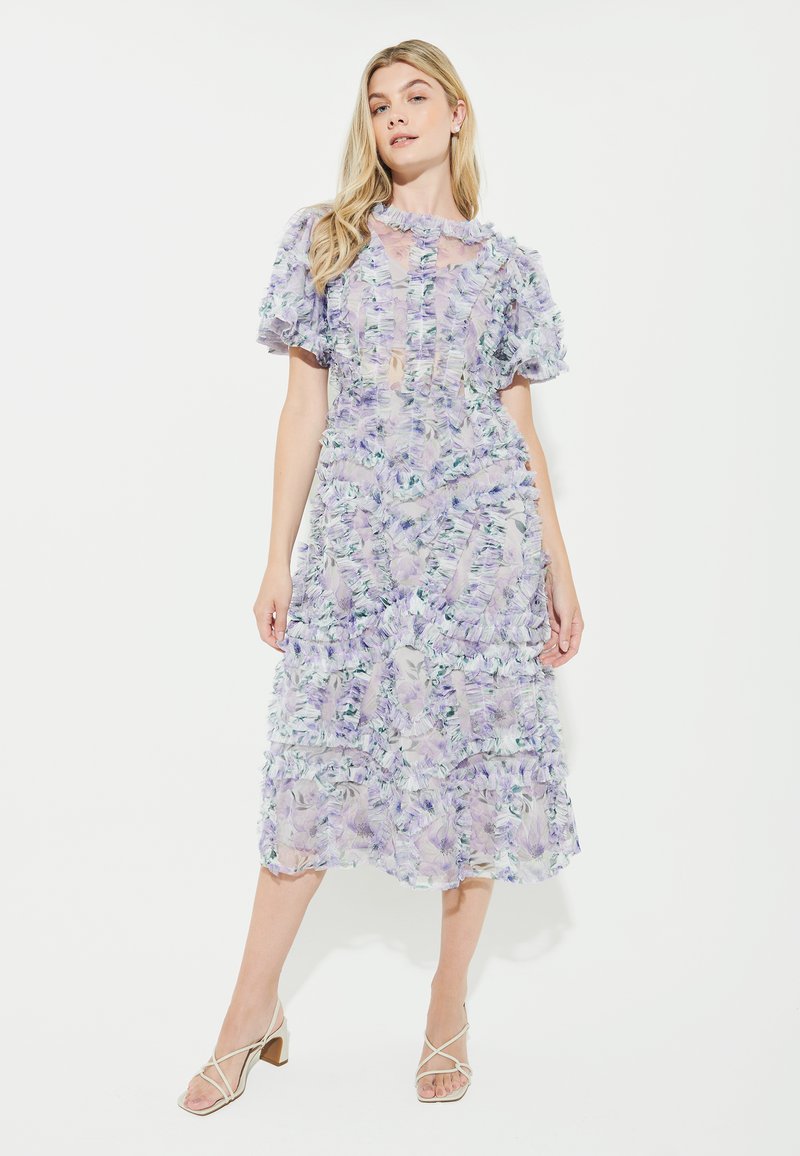 Lavendelfarbenes Blumenkleid mit Rüschenlagen und Puffärmeln, das aus einem durchsichtigen Stoff gefertigt ist und ein hochgeschlossenes Design aufweist. Beigefarbene, geschnürte Schuhe runden den Look ab.
