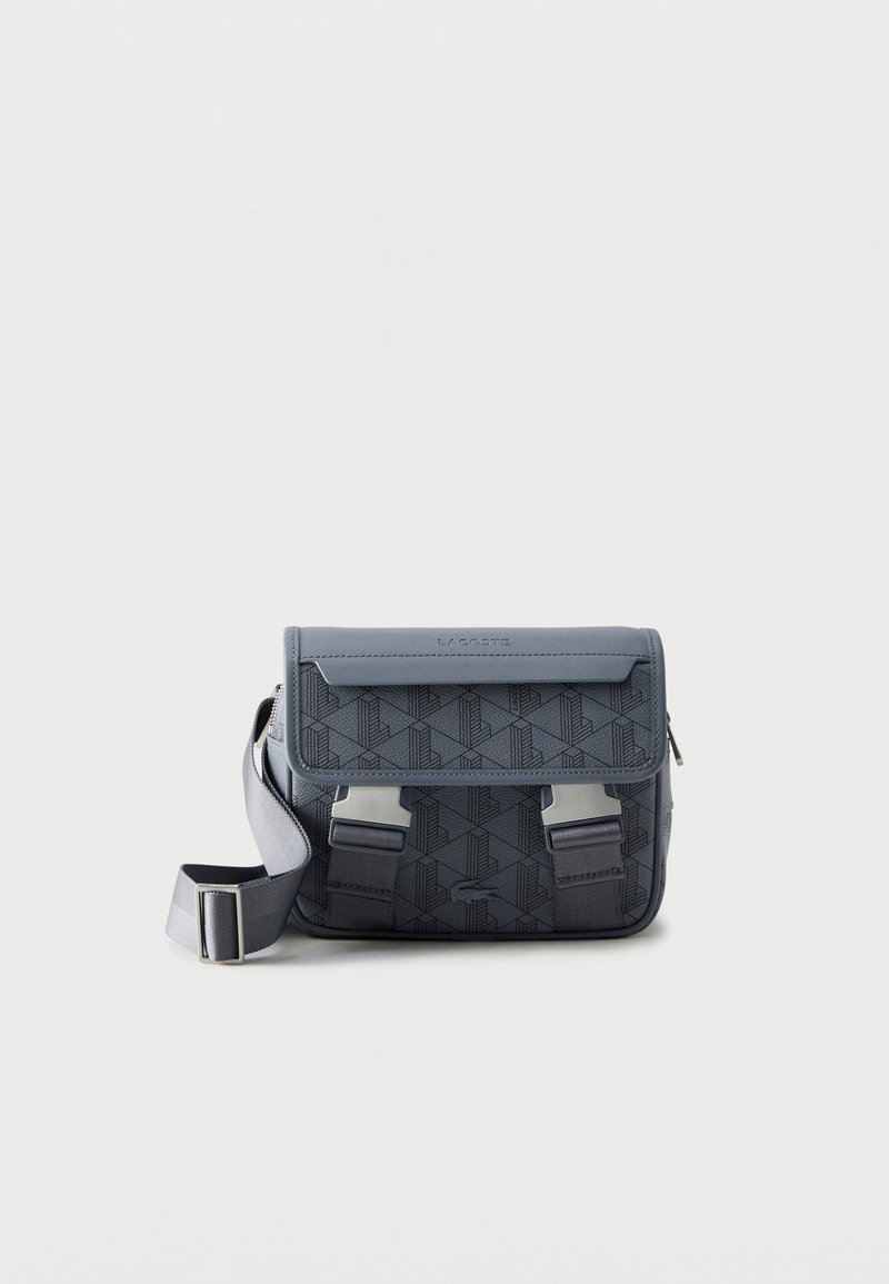 Σκούρο γκρι τσάντα χιαστί με γεωμετρικό μοτίβο, δύο ασημένιες αγκράφες μπροστά, ρυθμιζόμενο λουρί και λογότυπο Lacoste στο καπάκι.