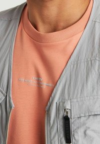 Giacca grigia chiaro con cerniera sopra una felpa color pesca. La felpa presenta un colletto a coste e dettagli testuali in bianco.