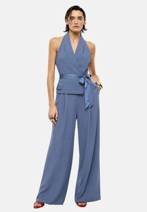 Donna che indossa un top halter blu senza maniche con cintura in vita e pantaloni a gamba larga abbinati, abbinati a scarpe rosse open toe e gioielli in oro.