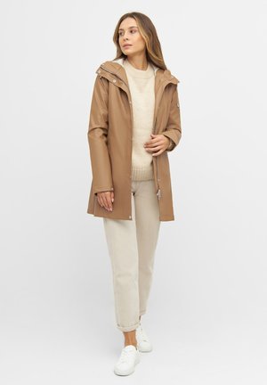 Chaqueta de lluvia impermeable marrón con capucha, cierre de cremallera y botones a presión. Llevada sobre un suéter beige y pantalones de color claro. Zapatillas blancas.
