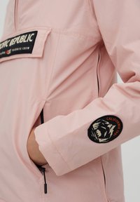 Lys pink jakke med en frontlomme, der har teksten "PACIFIC REPUBLIC." Venstre ærme viser et rundt mærke med et bjørne-design.