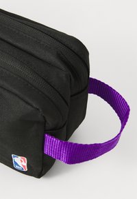 Sort stofpose med tekstureret overflade, lilla vævet håndtag og lille NBA-logo patch. Rektangulær form med dobbelte lynlåslukninger.