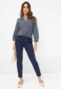 Marinebluse mit weißem kreisförmigem Muster, langen Ärmeln und V-Ausschnitt. Enganliegende marineblaue Hose und hellbeige Handtasche, dazu beige Ballerinas.