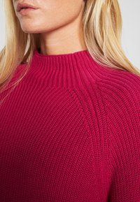 Fuksjowy sweter dziany z wysokim kołnierzem, z prążkowaną fakturą i ukośnymi przeszyciami na ramionach.