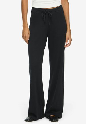 Femme portant un pantalon noir à jambes larges avec une taille à cordon, associé à des chaussures plates noires et un haut blanc, debout devant un fond uni.