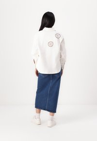 Cămașă albă cu nasturi, decorată cu două accente circulare din dantelă pe spate, asortată cu o fustă midi din denim albastru și adidași albi.