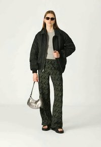 Giacca bomber nera, top grigio, pantaloni flared verdi a fantasia, sandali neri, borsa argento, occhiali da sole. Elementi di design lucidi e moderni.