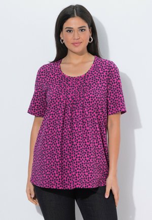 Frau mit langen dunklen Haaren, die silberne Creolen und eine rosa gemusterte Bluse mit kurzen Ärmeln trägt, steht vor einem weißen Hintergrund.