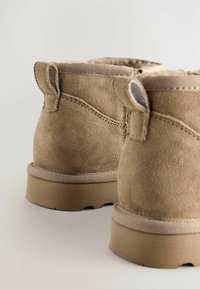 Botas de tobillo de ante marrón con una textura suave, costuras reforzadas, acentos de lona y lengüetas en el collar. Suelas de goma planas.