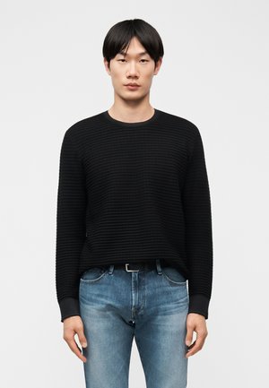 Ázijský muž stojaci, oblečený v čiernom rebrovanom svetri s dlhými rukávmi a svetlomodrých džínsoch s čiernym opaskom, otočený čelom na bielom pozadí.