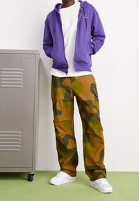 Sudadera morada con cremallera sobre una camiseta blanca, combinada con pantalones cargo de camuflaje en marrón y verde, y zapatillas deportivas blancas. Taquilla plateada cerca.