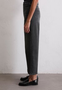 Szare dżinsy o wysokim stanie, proste nogawki, z bocznymi kieszeniami i jedną kieszenią z tyłu, zestawione z błyszczącymi czarnymi mokasynami.