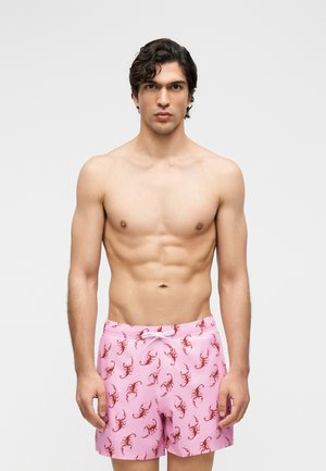 Oberkörperfreier Mann mit kurzen lockigen Haaren, der rosa Badeshorts mit roten Skorpionen trägt und vor einem einfarbigen Hintergrund steht.