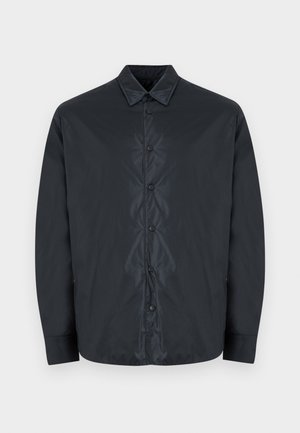 Chemise noire à manches longues avec un col pointu et cinq boutons-pression visibles au centre devant.