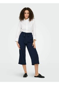 Chemise blanche à boutons avec manches longues associée à un pantalon large cropped bleu marine et des chaussures noires sans lacets. Design minimaliste et élégant.