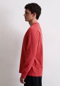 Langarm T-shirt in koraal, gemaakt van zachte katoen. Kenmerkt zich door een ronde halslijn en een relaxte pasvorm, gecombineerd met zwarte broeken. Zijaanzicht weergegeven.